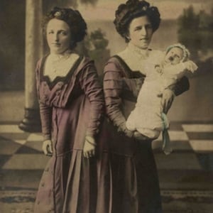 Op de afbeelding: Twee vrouwen in lange, donkerpaarse jurken staan ​​naast elkaar. De vrouw rechts houdt een baby vast in een witte jurk met een blauw lint. De vrouwen poseren in een studio met een geruite vloer en een geschilderde achtergrond.