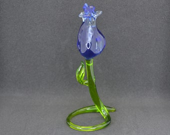 May include: A glass sculpture of a blue flower with a green stem and leaves. The stem is curled into a circle at the base.