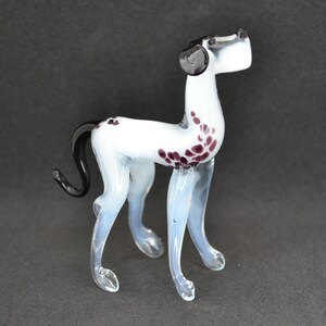 Op de afbeelding: Een wit glazen beeldje van een hond met paarse vlekken en een zwarte staart. De hond staat op alle vier poten en heeft zijn kop omhoog gekanteld.