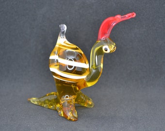 May include: A glass figurine of a yellow and white striped bird with a red beak and a long red neck. The bird is standing on a black surface.