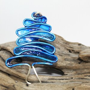 May include: A blue and silver metal Christmas tree bauble with a layered design. The bauble is sitting on a piece of driftwood.