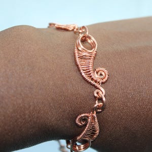 Op de afbeelding: Een koperdraad armband met ingewikkelde, met de hand gewikkelde metalen spoelen en spiraalvormige ontwerpen. De armband is opgebouwd uit meerdere onderling verbonden segmenten van gepolijst koperdraad met een gedetailleerd geweven patroon.