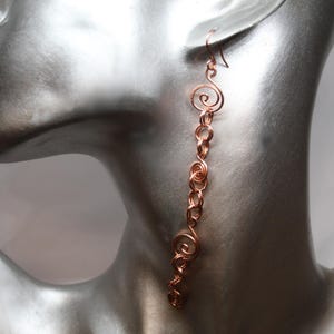 以下が含まれることがあります： らせん状のデザインが施された長い銅線製のイヤリングのペア。イヤリングはマネキンの耳からぶら下がっています。