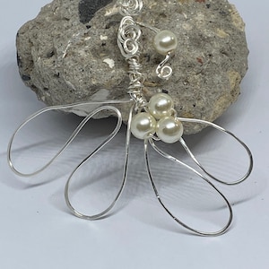 Op de afbeelding: Een zilveren draadbloem met witte parels. De bloem heeft vijf bloemblaadjes en een steel met een lus aan de bovenkant.