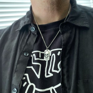 May include: A silver skull pendant necklace on a silver chain. The pendant is a detailed skull with a textured surface. The necklace is worn by a person wearing a black shirt with a white graphic design.