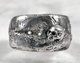May include: A silver ring with a textured surface and a small skull design. The ring has three small diamonds set into the surface.