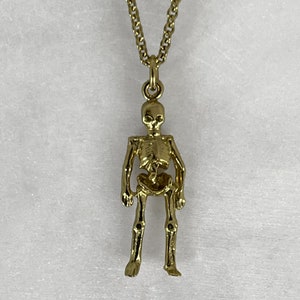 May include: A gold skeleton pendant necklace. The skeleton is standing with its arms at its sides. The necklace is on a white background.