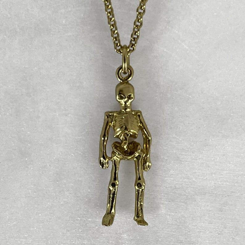 May include: A gold skeleton pendant necklace. The skeleton is standing with its arms at its sides. The necklace is on a white background.