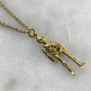 May include: A gold chain necklace with a detailed skeleton pendant. The skeleton is standing with its arms at its sides.
