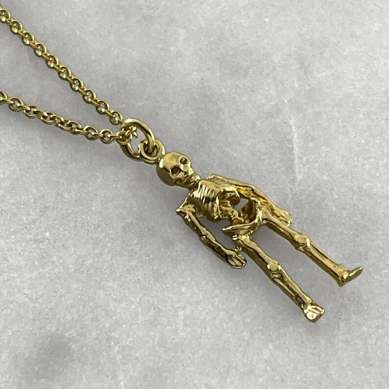 May include: A gold chain necklace with a detailed skeleton pendant. The skeleton is standing with its arms at its sides.