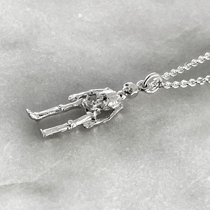 May include: A silver skeleton pendant necklace. The pendant is a detailed, three-dimensional skeleton figure with a chain link attached to the top of the skull.