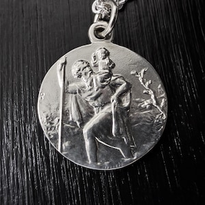 Könnte beinhalten: Ein runder silberfarbener Anhänger mit einem erhabenen Reliefdesign, das eine Figur zeigt, die ein kleines Kind auf der Schulter trägt und einen langen Stab hält. Der Anhänger ist oben mit einer runden Öse an einer silberfarbenen Kette befestigt.