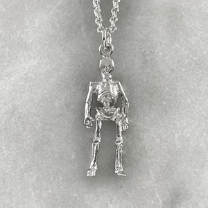 May include: Silver skeleton pendant necklace. The skeleton is standing with its arms at its sides. The necklace is on a white background.