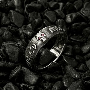 May include: A silver ring featuring a skull design with the inscription "Memento Mori" around the band. The skull has red eyes.