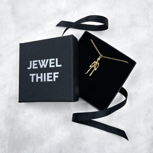 May include: A gold skeleton pendant necklace in a black box with a black ribbon. The box has the words "JEWEL THIEF" printed on the front.