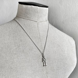 May include: A silver chain necklace with a small silver figure pendant. The figure is standing with its arms at its sides.