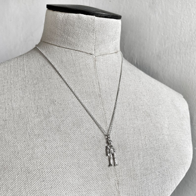 May include: A silver chain necklace with a small silver figure pendant. The figure is standing with its arms at its sides.