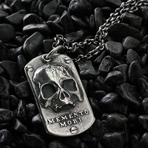 May include: A silver dog tag necklace featuring a skull design and the inscription "Memento Mori".