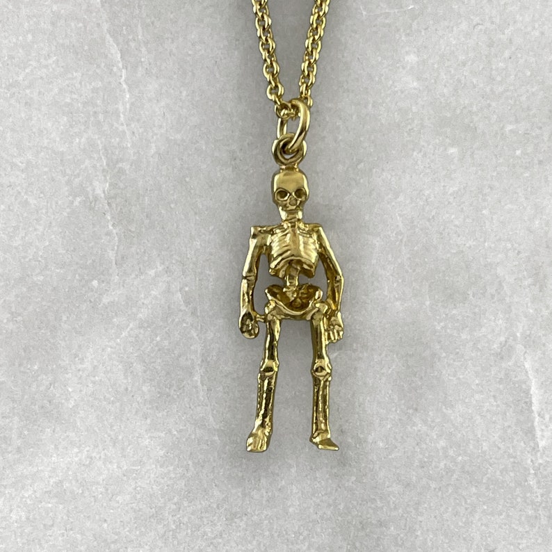 May include: A gold skeleton pendant necklace. The skeleton is standing with its arms at its sides. The necklace is on a white background.