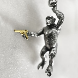 May include: A silver monkey figurine holding a gold-colored gun. The monkey is hanging from a chain and has a textured surface.