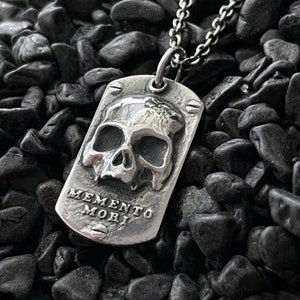 May include: A silver pendant necklace featuring a skull design and the inscription "Memento Mori" on the front. The pendant is suspended from a silver chain.