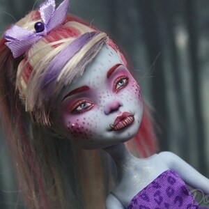 May include: A blue-skinned doll with pink and white hair styled in a ponytail. The doll has blue eyes, pink eyeshadow, and a purple and white bow in her hair. She is wearing a purple and white patterned top.