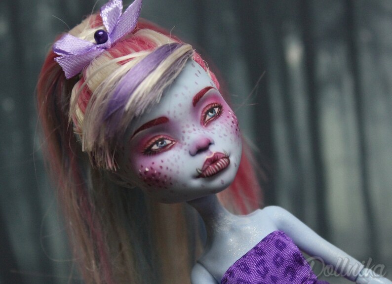 May include: A blue-skinned doll with pink and white hair styled in a ponytail. The doll has blue eyes, pink eyeshadow, and a purple and white bow in her hair. She is wearing a purple and white patterned top.