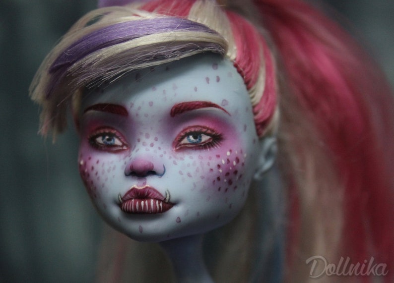 May include: Close-up of a doll's face with blue skin, pink and white makeup, and pink and white hair. The doll has a unique, slightly scary expression.