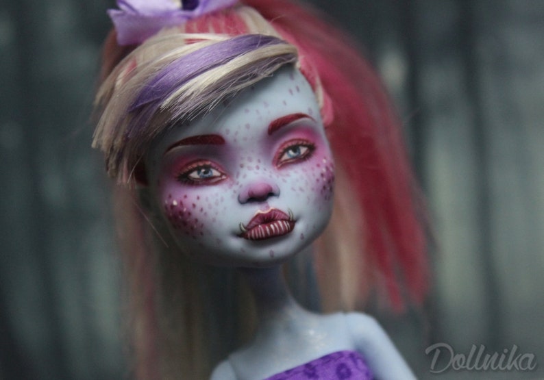 May include: A close-up of a doll's face with blue skin, pink and white hair, and purple eyeshadow. The doll has a unique, slightly scary expression with a wide smile and sharp teeth.