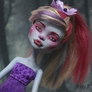 May include: A custom-made doll with blue skin, pink and white hair, and purple clothing. The doll has a painted face with pink blush and blue eyes.