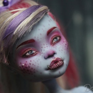 May include: A close-up of a doll's face with blue skin, pink and red makeup, and a purple and white wig. The doll has a pink and white bow in her hair and a small, silver nose ring.