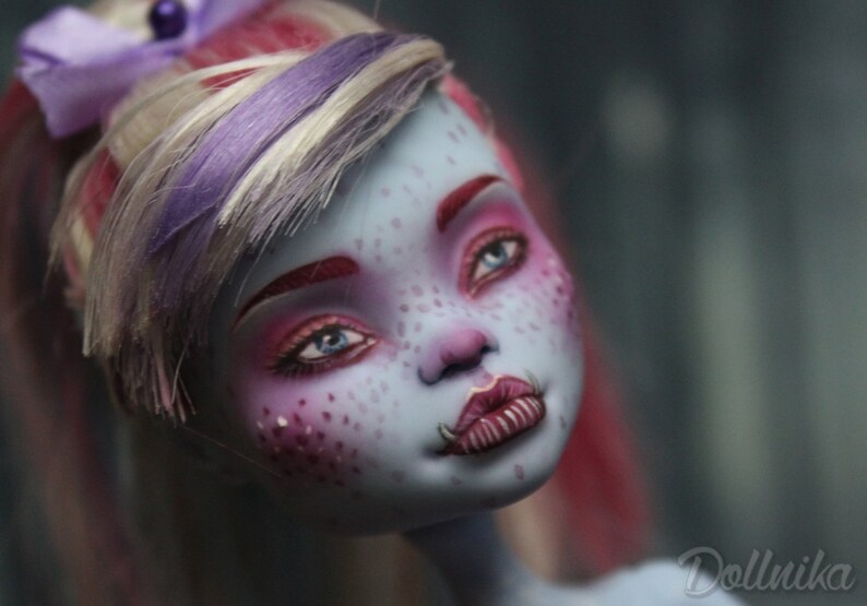 May include: A close-up of a doll's face with blue skin, pink and red makeup, and a purple and white wig. The doll has a pink and white bow in her hair and a small, silver nose ring.
