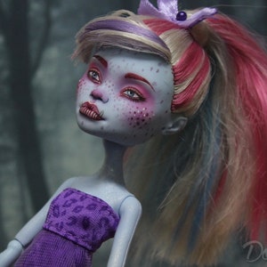 May include: A blue-skinned doll with pink, blonde, and blue hair styled in a ponytail. The doll is wearing a purple strapless dress with a leopard print pattern. The doll has blue eyes and pink eyeshadow.