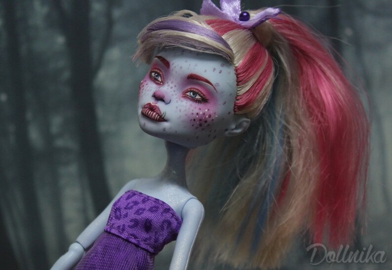 May include: A blue-skinned doll with pink, blonde, and blue hair styled in a ponytail. The doll is wearing a purple strapless dress with a leopard print pattern. The doll has blue eyes and pink eyeshadow.