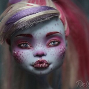 May include: A close-up of a doll's face with blue skin, pink and purple hair, and bright pink and blue eye makeup. The doll has a freckled face and a toothy grin.