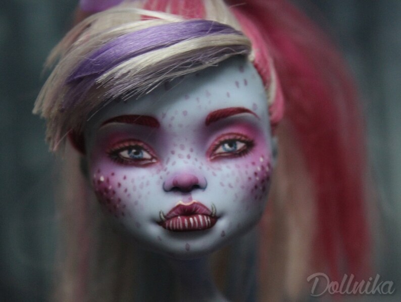 May include: A close-up of a doll's face with blue skin, pink and purple hair, and bright pink and blue eye makeup. The doll has a freckled face and a toothy grin.