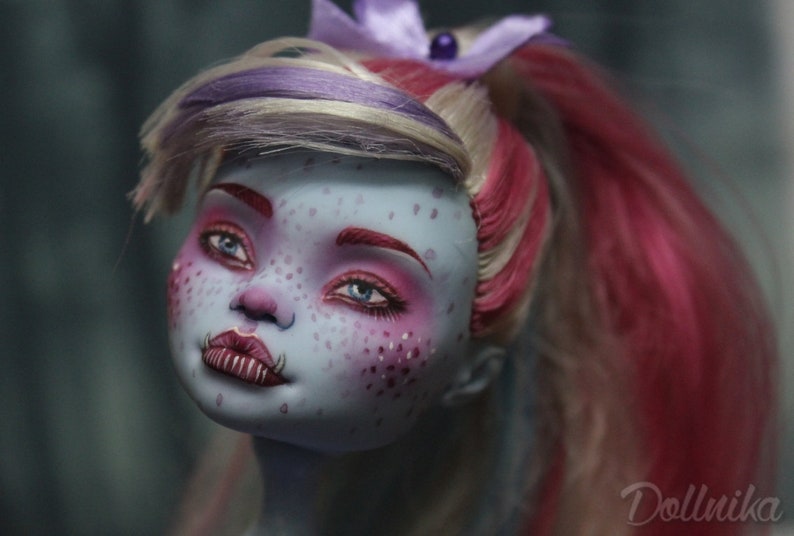 May include: A blue-skinned doll with pink and white hair, wearing pink eyeshadow and lipstick. The doll has a nose ring and fangs.