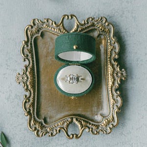 May include: An engagement ring with three stones rests in an open, green velvet ring box. The ring box sits on a gold-colored, ornate tray. A wooden object and greenery are also in the frame.