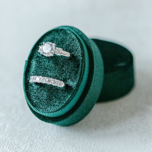 May include: Two silver rings with diamonds are displayed in a green velvet ring box. The box is round and has a lid. The lid is open and the rings are visible. The box is on a white surface. BOX COLOR: EMERALD