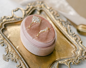 May include: Two gold rings with diamonds are displayed in a pink velvet ring box. The box is sitting on a gold ornate tray.