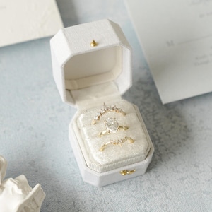 May include: A white, octagonal ring box with a gold clasp and button, open to reveal three gold rings with gemstones. The rings are set in a white velvet interior. The background includes wedding stationery and a small white bust.