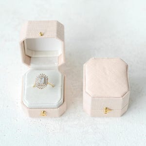 May include: Two light pink octagonal ring boxes, one open and one closed. The open box displays a gold ring with a square-cut center stone surrounded by smaller stones. The boxes have gold-colored clasps.