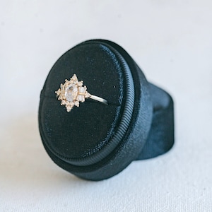 May include: A gold ring with a large oval-shaped clear stone surrounded by smaller clear stones in a geometric pattern. The ring is displayed on a black velvet ring box.