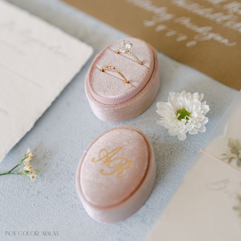 May include: Two engagement rings in a mauve velvet ring box with the initials "A & R" on the lid. The box is on a light blue surface.