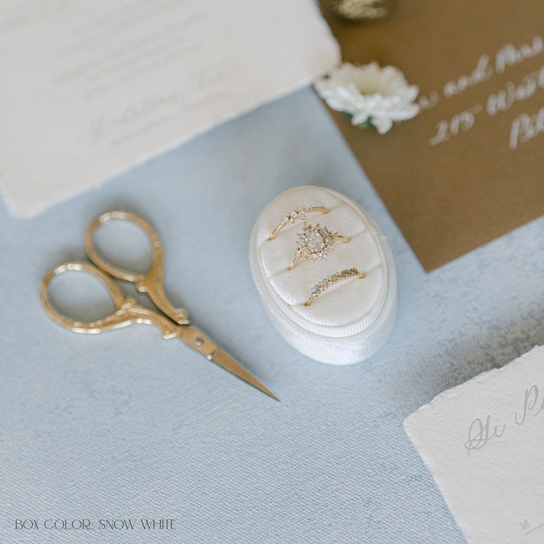 May include: A white velvet ring box with four gold rings, including a large center ring with a diamond, on a light blue surface. A pair of gold scissors is also on the surface.