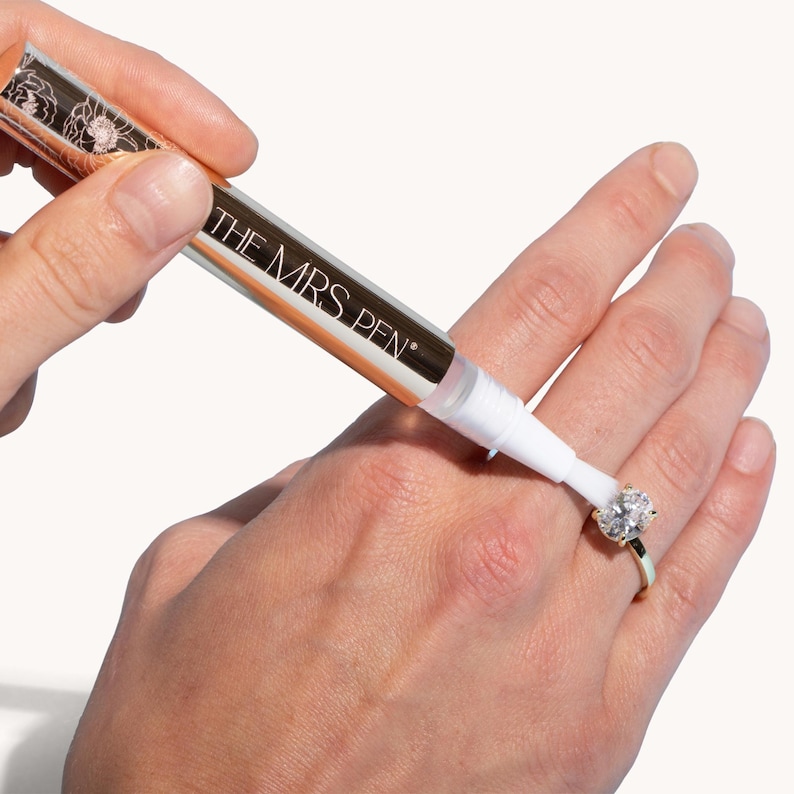 K&ouml;nnte beinhalten: Ein goldener und wei&szlig;er Stift mit der Aufschrift "THE MRS. PEN" wird verwendet, um einen Diamantring an einem Finger zu reinigen.