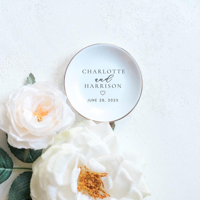 May include: A white ceramic ring dish with gold trim, featuring the names "Charlotte and Harrison", a heart symbol, and the date "June 28, 2025". The dish is surrounded by white roses.