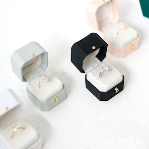 May include: A selection of open ring boxes in black, pink, and green, each displaying an engagement ring. The boxes have a velvet interior and a gold clasp. The rings feature different diamond cuts, showcasing the jewellery.