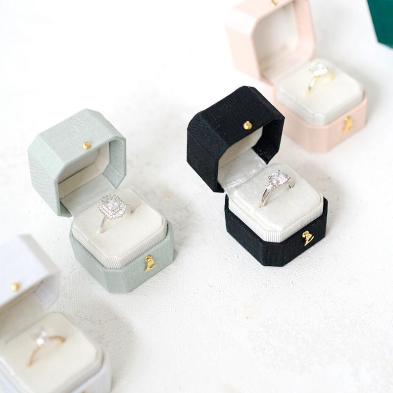 May include: A selection of open ring boxes in black, pink, and green, each displaying an engagement ring. The boxes have a velvet interior and a gold clasp. The rings feature different diamond cuts, showcasing the jewellery.