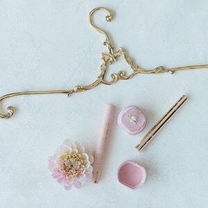 K&ouml;nnte beinhalten: Ein goldener, verzierter Kleiderb&uuml;gel mit einer rosafarbenen Ringbox, einer rosafarbenen Ringbox mit einem Diamantring, einer rosafarbenen Lippenbalsamtube, einer rosafarbenen Blume und einem goldenen Eyeliner-Stift.